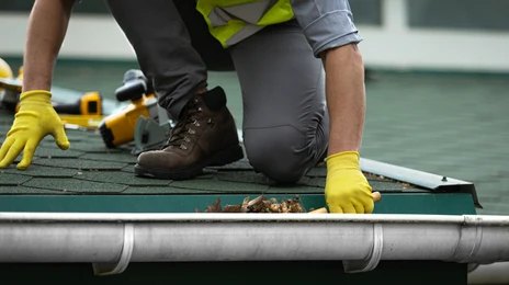A person wearing gloves cleans debris from a rooftop rain gutter.